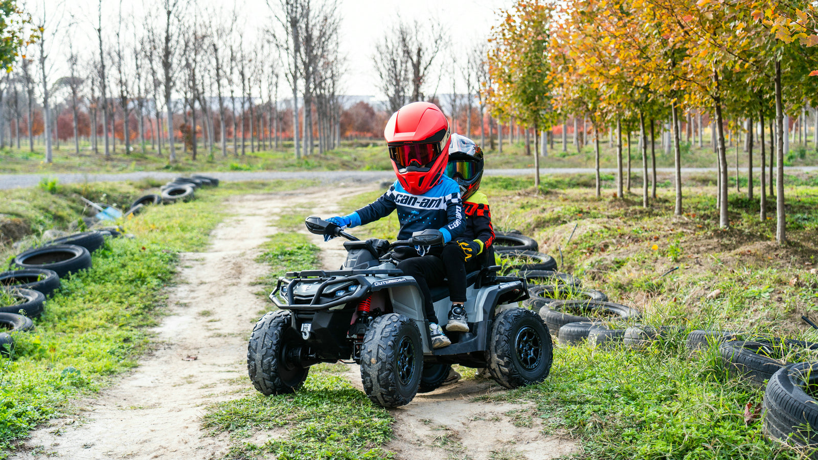 Two children wearing helmets ride a black Can-Am Outlander kids ATV along a dirt track lined with trees.