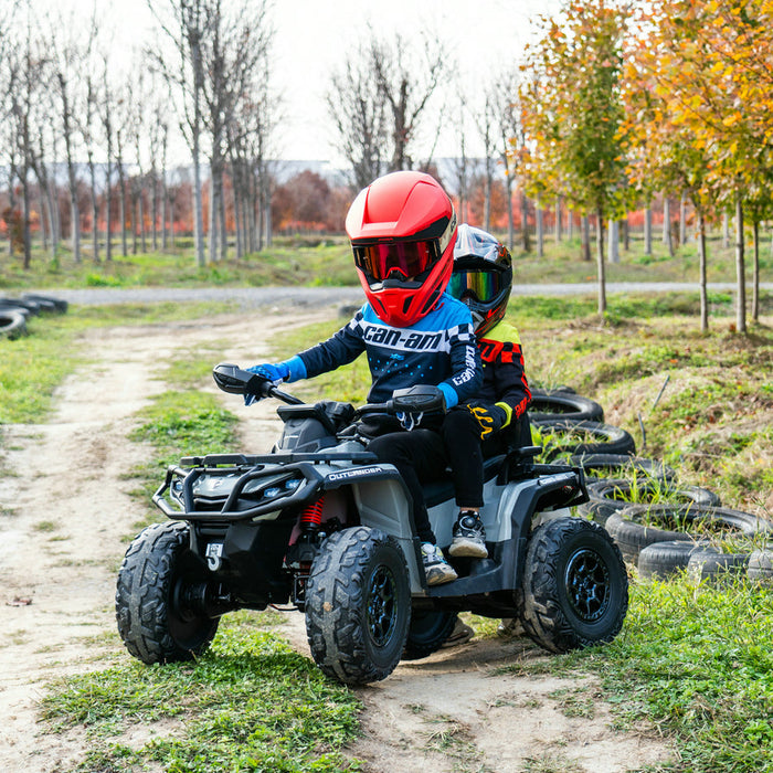 Two children wearing helmets ride a black Can-Am Outlander kids ATV along a dirt track lined with trees.