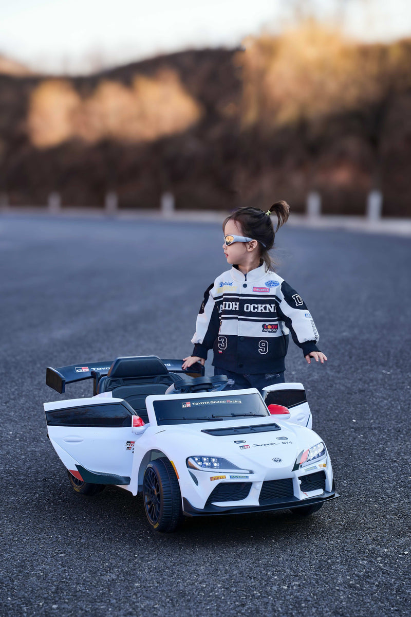 A young child wearing a black and white racing jersey stands behind a white and black Toyota Supra ride-on race car on an asphalt road with a blurred outdoor background.