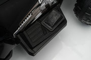 Close-up detail shot of a black electric ride-on toy vehicle, showing the side step platform, textured grip pad, and part of the treaded rear wheel on a white studio background.